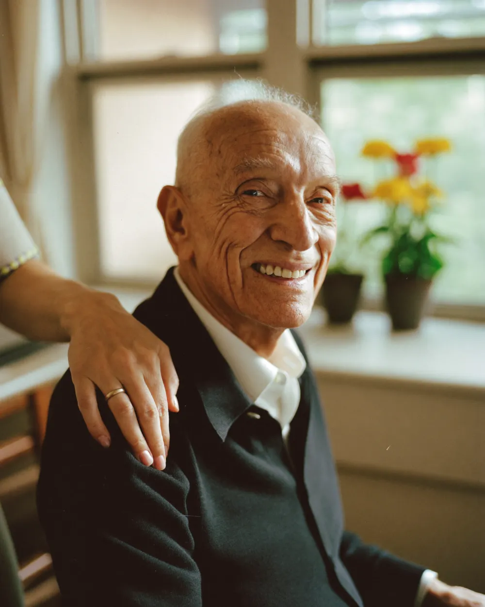 An older man smiling warmly with a caregiver’s hand on his shoulder